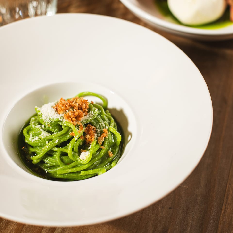 Green pasta topped with grated cheese and toasted breadcrumbs on a white plate, with another plated dish and water glass in the background.
