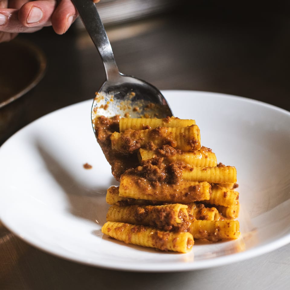 Hand serves rigatoni pasta coated in rich meat sauce onto a white plate.
