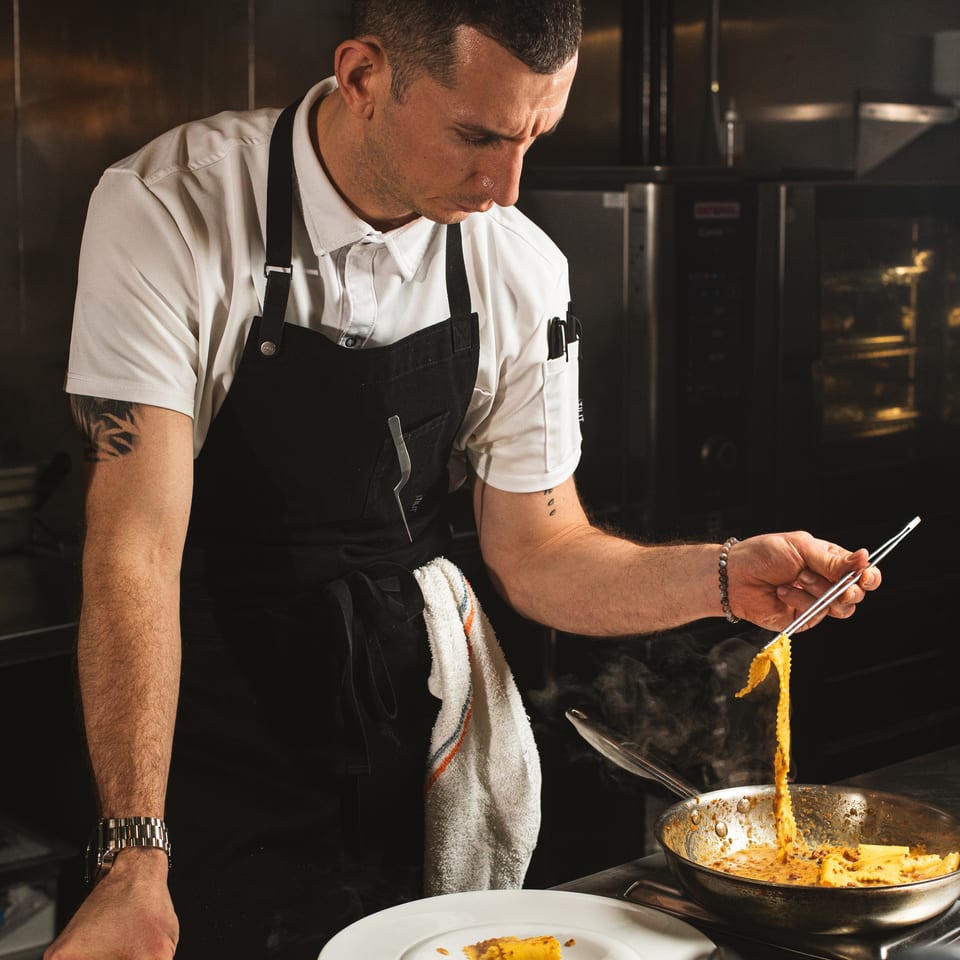 Chef in a black apron plates steaming pasta from a skillet in a dimly lit restaurant kitchen.