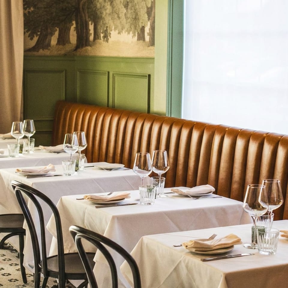 Restaurant dining room with white-clothed tables, black chairs, a brown leather banquette, and green paneled walls.