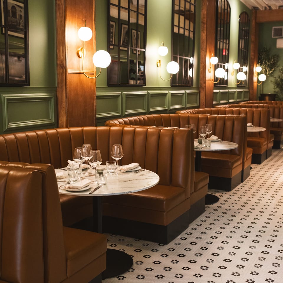 Elegant restaurant dining room with brown leather booths, marble tables, globe sconces, green walls, wood trim, and patterned tile flooring.