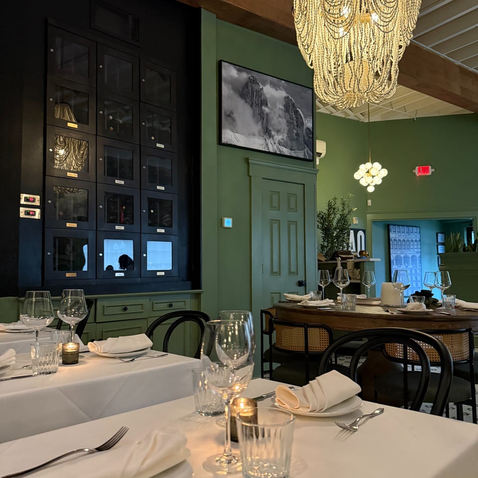 Elegant restaurant dining room with white-tablecloth tables, wine glasses, green walls, and a large illuminated beaded chandelier.