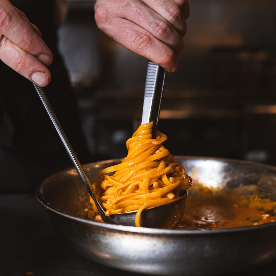 Chef twirls glossy orange-sauced noodles with metal tongs into a black bowl over a stainless-steel pan.