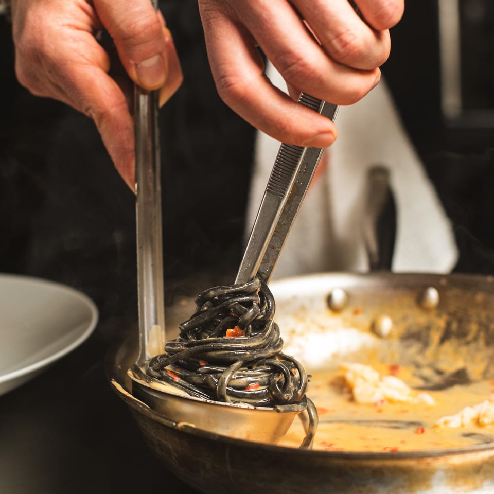 Chef uses tongs to arrange black pasta in a bowl beside a large serving platter.