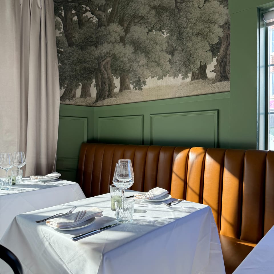 Elegant restaurant dining area with white tablecloths, set glassware, caramel leather booths, green paneling, and forest mural wallpaper.